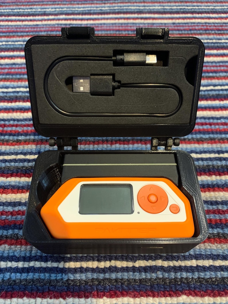 Puede incluir: Una caja de transporte negra con un peque&ntilde;o dispositivo naranja y blanco en su interior. El dispositivo tiene una pantalla y botones. Se incluye un cable USB negro.