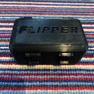 Puede incluir: Una caja de pl&aacute;stico negra con la palabra "FLIPPER" impresa en la parte superior.