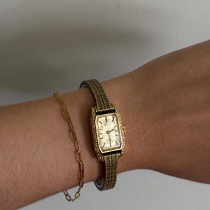 Puede incluir: Un reloj de pulsera rectangular en tono dorado con una correa de metal texturizada y una pulsera de eslabones a juego. El reloj presenta una esfera rectangular de color claro con índices finos y una pequeña corona lateral.