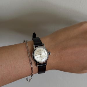 Könnte beinhalten: Eine silberfarbene analoge Armbanduhr mit rundem weißem Zifferblatt, schwarzen arabischen Ziffern und einem schwarzen Lederarmband. Die Uhr trägt den Markennamen Timex auf dem Zifferblatt. Ein zartes silberfarbenes Kettenarmband mit länglichen ovalen Gliedern wird dazu getragen.