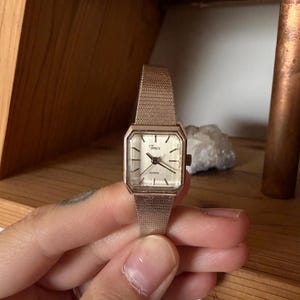 May include: A vintage Timex quartz watch with a rectangular gold-toned face and a mesh strap. The watch face is light-coloured with black hands and markers. The strap is a matching gold colour. The watch is a classic style.