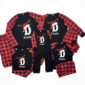Pode incluir: Pijamas de Natal familiares a condizer em xadrez preto e vermelho. As partes de cima pretas apresentam um gorro de Pai Natal, chifres e a letra "D" com o nome "Dustin". Inclui camisas de manga comprida, calças e um body.