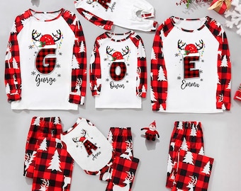 Op de afbeelding: Bijpassende kerstpyjama's voor het gezin in rood en zwart buffelruit. De witte tops hebben rode mouwen en een rendierontwerp met een letter en naam. De pyjamabroeken zijn rood en zwart geruit met witte bomen en rendieren.