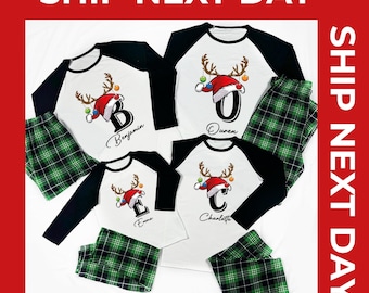 Op de afbeelding: Wit en zwart raglanmouw pyjama met een kerstthema. Elk shirt heeft een letter en naam, met een kerstmuts en rendiergeweien ontwerp. De bijpassende groene geruite broek maakt de set compleet. De tekst "SHIP NEXT DAY" wordt weergegeven.