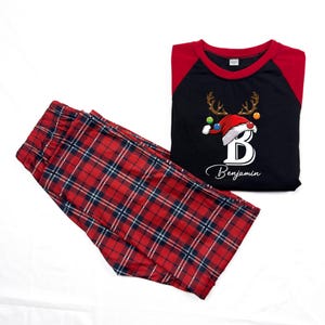 K&ouml;nnte beinhalten: Rote und schwarze karierte Schlafanzughose mit einem schwarzen Langarmshirt mit einem Rentier mit Weihnachtsm&uuml;tze und dem Buchstaben "B" mit dem Namen "Benjamin" darunter.