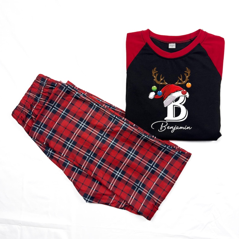 K&ouml;nnte beinhalten: Rote und schwarze karierte Schlafanzughose mit einem schwarzen Langarmshirt mit einem Rentier mit Weihnachtsm&uuml;tze und dem Buchstaben "B" mit dem Namen "Benjamin" darunter.