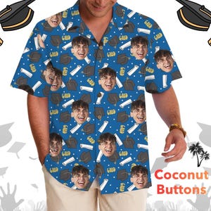 May include: Blue button-up shirt with a repeating pattern of graduation caps, smiling faces, and the word "GRAD". The shirt has coconut buttons and short sleeves. The shirt is worn by a person.