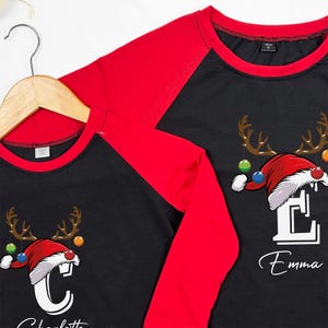 K&ouml;nnte beinhalten: Zwei lang&auml;rmelige Shirts in Schwarz und Rot mit einem Weihnachts-Rentier-Design. Die Shirts zeigen einen wei&szlig;en Buchstaben, "C" und "E", mit einem Weihnachtsmann-Hut und Rentiergeweih. Die Shirts haben die Namen "Charlotte" und "Emma" unter den Buchstaben gedruckt.