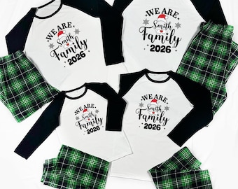 May include: A set of four matching pajama outfits featuring white long-sleeve shirts with black raglan sleeves and green and black plaid pants. The shirts display the text "WE ARE Smith Family 2026" with a small red and white Santa hat graphic.