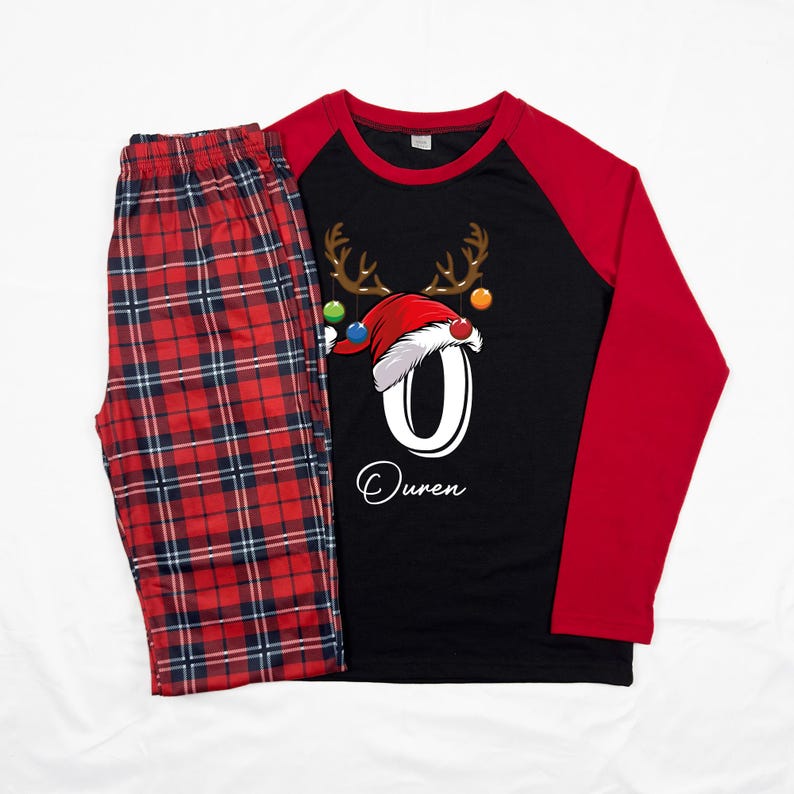 K&ouml;nnte beinhalten: Rote und schwarze karierte Schlafanzughose mit einem schwarzen Langarmshirt mit einem Rentier mit einem Weihnachtsmann-Hut und der Zahl 0. Das Shirt hat rote &Auml;rmel und der Name "Ouren" ist unter der Zahl gedruckt.