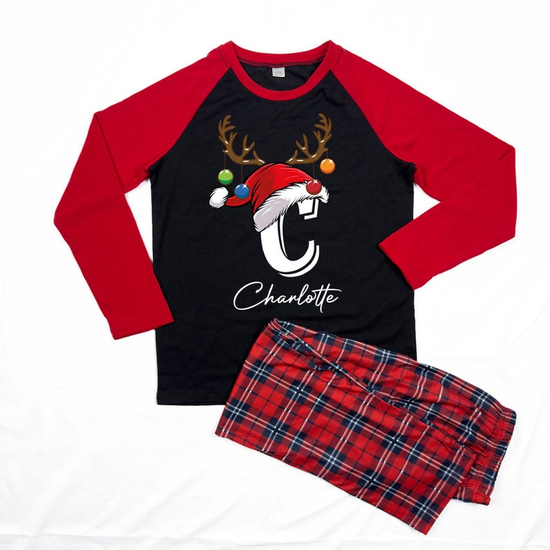 K&ouml;nnte beinhalten: Schwarz-roter lang&auml;rmliger Schlafanzug mit einem Rentier-Motiv und dem Buchstaben "C" in einer Weihnachtsm&uuml;tze. Der Schlafanzug hat ein Karomuster auf der Hose und der Text "Charlotte" ist auf dem Oberteil aufgedruckt.
