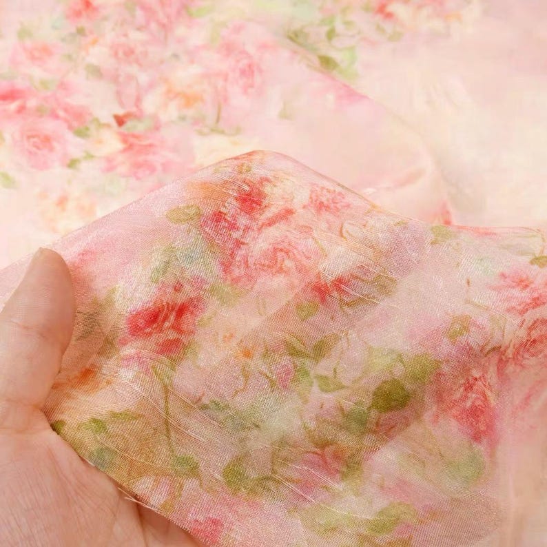 Op de afbeelding: Een delicate, doorschijnende stof in lichtroze, versierd met een bloemenprint. Het patroon toont roze, rode en groene bloemen. Het materiaal is doorschijnend, waardoor het een zachte, zachte uitstraling heeft, geschikt voor kleding of knutselprojecten.