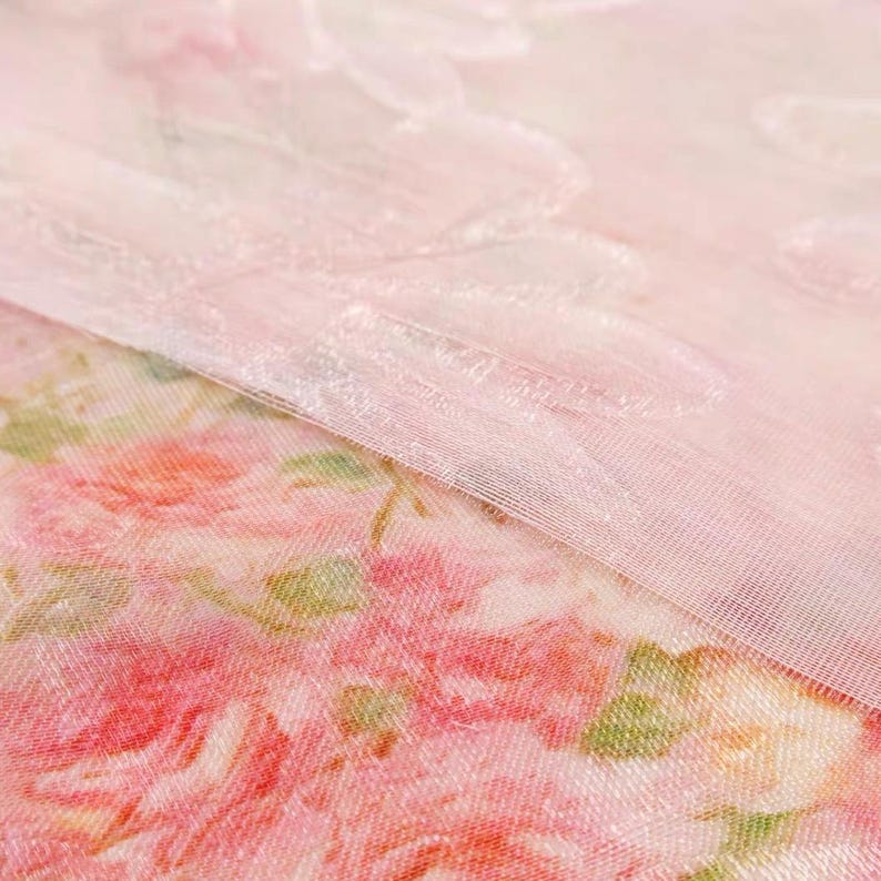 Op de afbeelding: Close-up van twee lagen stof. De onderste laag heeft een bloemenpatroon met roze en rode rozen en groene bladeren. De bovenste laag is een transparante, lichtroze stof met een subtiel, gestructureerd ontwerp. De stoffen overlappen elkaar.