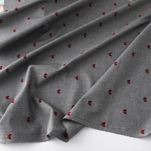 May include: A close-up shot of grey fabric with a repeating pattern of small, red floral designs. The fabric is draped, showing folds and texture. The red flowers are evenly spaced across the material.