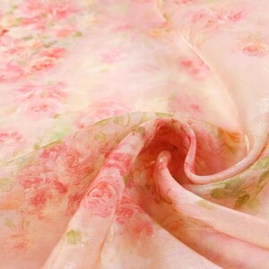 Op de afbeelding: Close-up van een delicate stof met een bloemenpatroon. De stof is zachtroze van kleur, met een herhalend patroon van roze en rode rozen en groene bladeren. De stof is licht doorschijnend en heeft een vloeiende, gedrapeerde uitstraling.