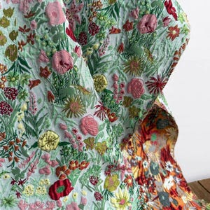 Puede incluir: Una tela con un patrón floral denso y texturizado sobre un fondo azul claro. El diseño incluye flores de estilo bordado en tonos rosa, rojo, amarillo y verde. Una sección de la tela se pliega para revelar una parte inferior en tonos naranja.