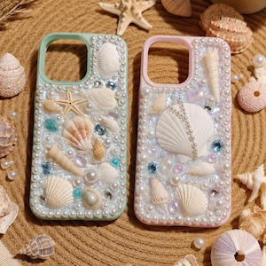 May include: Two phone cases, one mint green and one pink, are adorned with seashells, pearls, and crystals. The cases are surrounded by various seashells and a starfish on a textured, tan surface. The cases are designed to protect the phone.