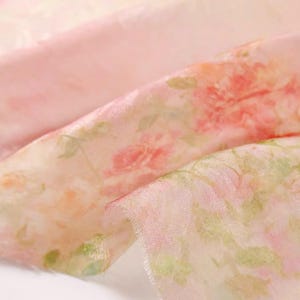 Op de afbeelding: Close-up van een delicate, transparante stof met een bloemenpatroon. De stof is voornamelijk lichtroze met een aquarelontwerp van rode en groene bloemen. Het materiaal lijkt zacht en vloeiend.