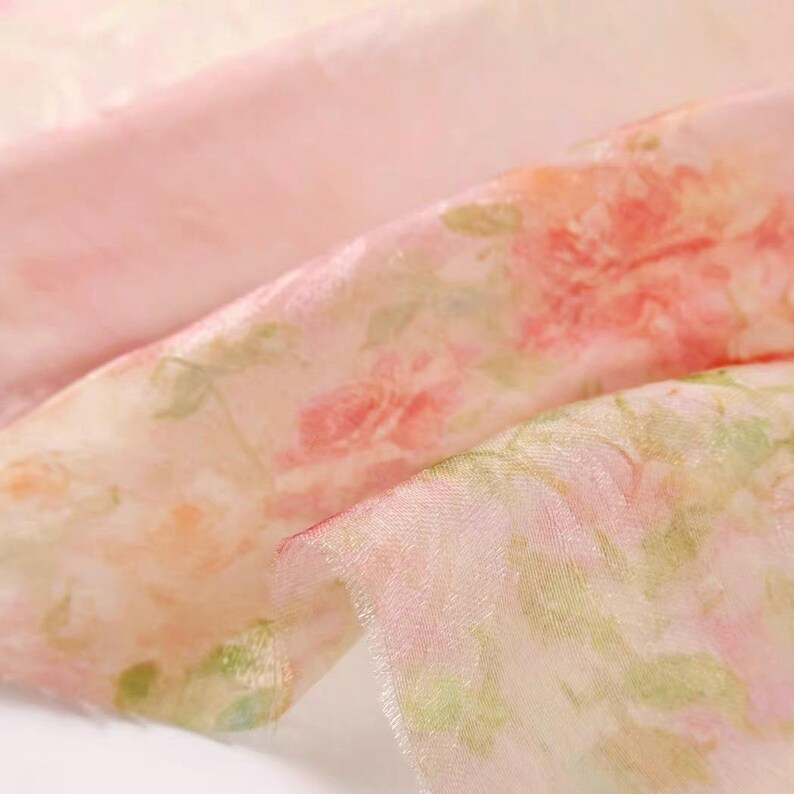Op de afbeelding: Close-up van een delicate, transparante stof met een bloemenpatroon. De stof is voornamelijk lichtroze met een aquarelontwerp van rode en groene bloemen. Het materiaal lijkt zacht en vloeiend.