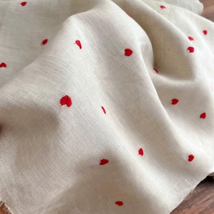 Könnte beinhalten: Nahaufnahme eines beigen Stoffes mit einem Muster aus kleinen, roten, gestickten Herzen. Der Stoff ist drapiert und zeigt seine Textur und das verstreute Herzdesign. Die Kanten des Stoffes sind ausgefranst.