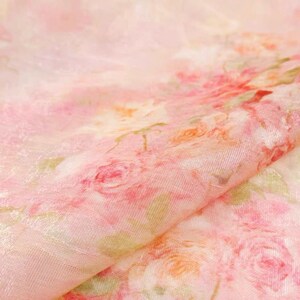Op de afbeelding: Close-up van een delicate stof met een bloemenpatroon. De stof heeft een zachtroze basis met rozen in tinten roze, rood en oranje. De stof heeft een subtiele glans, wat wijst op zijde of satijn. Het geheel is zacht en romantisch.