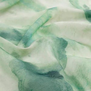 May include: Close-up of a flowing fabric with a watercolor effect. The fabric features a blend of white and shades of green, creating a soft, ethereal look. The design resembles abstract floral patterns, with the fabric's folds adding depth.