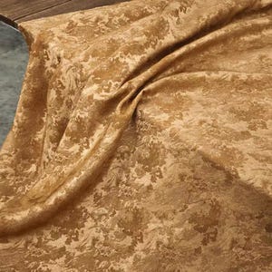 Könnte beinhalten: Ein Stoffstück in einem warmen goldbraunen Farbton mit einem tonalen, komplizierten floralen Damastmuster. Das Material ist drapiert, um weiche Falten und einen subtilen Glanz auf der strukturierten Oberfläche zu zeigen, was die detaillierten botanischen Motive hervorhebt.