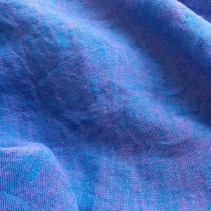 May include: A piece of woven fabric featuring a vibrant blend of blue and purple threads. The material displays a distinct textured surface with visible cross-hatching patterns created by the interlacing of the colored fibers throughout the textile.