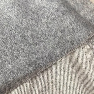 Könnte beinhalten: Eine Nahaufnahme von zwei verschiedenen Stofftexturen, die übereinander liegen. Die obere Schicht ist ein weiches, flauschiges, mittelgraues Filzmaterial mit dichtem Flor. Darunter liegt ein hellerer, beigefarbener Stoff mit sichtbarer Köperbindung.