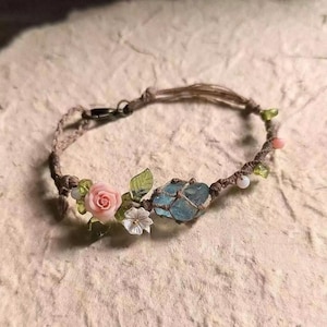 Peut inclure: Un bracelet fait à la main avec un cristal bleu clair enveloppé dans de la ficelle marron. Le bracelet est orné de petites roses roses, de fleurs blanches, de feuilles vertes et de petites perles. Le fermoir est en métal foncé.
