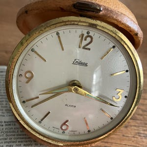 May include: A round Endura alarm clock with a gold-tone metal case and a tan leather-textured protective cover. The white dial features gold-tone numerals at 3, 6, 9, and 12, gold hands with light accents, and the text ALARM and WESTERN GERMANY.