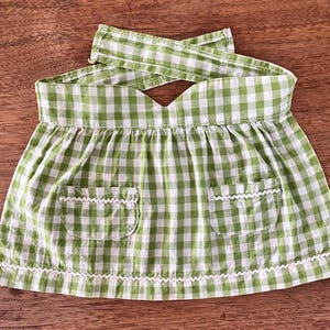 May include: A green and white gingham apron featuring a gathered skirt, two front pockets, and a waistband with crisscross straps. The edges of the pockets and the bottom hem of the apron are decorated with a thin, white, wavy rickrack trim.