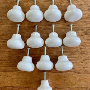 May include: A collection of twelve white ceramic cabinet knobs with metal screws. The knobs have a rounded, pear-like shape and are arranged on a wooden surface. Ideal for furniture restoration or DIY projects.