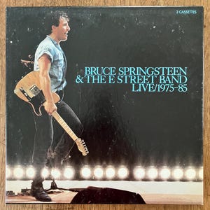May include: A square music album cover featuring Bruce Springsteen playing a light-colored electric guitar on stage. The text reads BRUCE SPRINGSTEEN & THE E STREET BAND LIVE/1975-85 and 3 CASSETTES. The background is black with a row of stage lights.