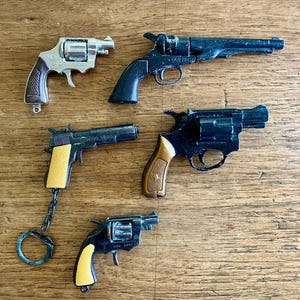 May include: Five toy guns of various sizes and designs are arranged on a wooden surface. The guns are primarily black and silver, with some featuring yellow or brown grips. One gun is attached to a keychain.