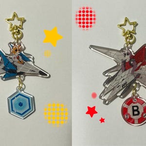 May include: Two clear acrylic keychains featuring anime characters in fighter jets. The keychain on the left features a blue and white jet with a character in a yellow and white pilot suit. The keychain on the right features a white and grey jet with a character in a red and white pilot suit. Both keychains have a star-shaped charm at the top and a charm with a letter on the bottom.