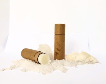 May include: Two brown cardboard tubes containing white lip balm. The tubes are on a white surface with white salt crystals.