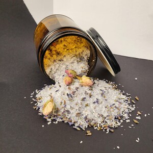 May include: A brown glass jar with a black lid filled with white bath salts and dried lavender and rose petals. The jar is tipped over and the salts are spilling out onto a black surface.