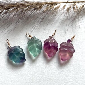 Peut inclure: Quatre pendentifs en forme de feuille dans des tons de bleu, vert, violet et rose. Chaque pendentif est fait d'un matériau translucide et possède une boucle en fil d'or sur le dessus. Les pendentifs sont disposés sur une surface blanche.