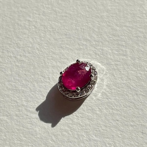 Peut inclure: Un pendentif argenté orné d'un rubis ovale, entouré de petits diamants transparents. Le rubis est d'un rouge profond et les pierres environnantes scintillent. Le pendentif est posé sur un fond blanc. Le design est élégant et attrayant.