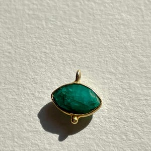 Può includere: Un ciondolo color oro con una pietra verde smeraldo a forma di goccia. La pietra è incastonata in una lunetta dorata con un piccolo anello nella parte superiore per una catena e una piccola perla decorativa nella parte inferiore.
