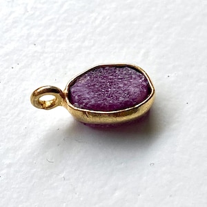 Può includere: Un piccolo ciondolo color oro con una gemma grezza di forma ovale e di colore viola intenso. La gemma è incastonata in una lunetta dorata e ha un piccolo anello per una catena. Il ciondolo è su sfondo bianco.