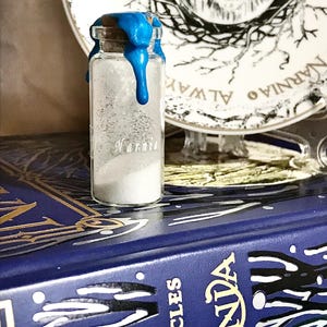 Könnte beinhalten: Eine kleine Glasflasche mit weißem Pulver darin. Die Flasche ist mit einem blauen Wachssiegel verschlossen. Die Flasche steht auf einem Buch mit dem Titel "Die Chroniken von Narnia".