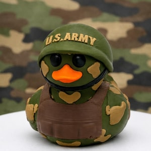 Peut inclure: Une figurine de canard en caoutchouc avec un motif camouflage vert et beige, un bec orange vif et des yeux noirs. Le canard porte un casque vert avec le texte U.S. ARMY en relief et un gilet tactique marron sur la poitrine.