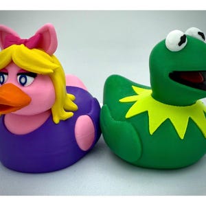 May include: Two novelty duck figurines. One is a pink duck with a yellow wig, a pink bow, and a purple dress. The other is a green frog-like duck with a yellow collar. Both are on a white surface.