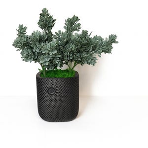 May include: A small artificial green plant with textured, sage-colored leaves arranged in a dense cluster. The plant is potted in a black, cylindrical container featuring a cross-hatched, diamond-patterned exterior and a small circular detail.