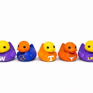 Op de afbeelding: Vijf rubberen eendjes op een rij, elk met een ander kleurenschema en universiteitsteamlogo. De eenden hebben gele koppen, oranje of paarse lichamen en teamlogo's zoals "W", "Boise State", "T", "Texas" en "LSU".