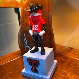 May include: A 3D-printed figurine of a cowboy with a red beard, wearing a red shirt with the number 12, a black hat, and black pants. The figurine stands on a light blue pedestal with a red and black logo.