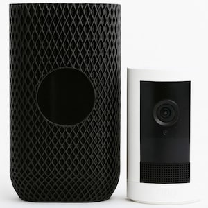 Puede incluir: Un objeto cilíndrico negro con un exterior en forma de diamante y una abertura circular, junto a una cámara de seguridad blanca y negra. La cámara tiene una lente y orificios para el altavoz.