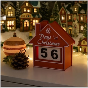 May include: An orange house-shaped Christmas countdown calendar showing "Days 'til Christmas" and the number "56". A decorative bauble and pine cone are in front of the calendar. Miniature illuminated houses are in the background.